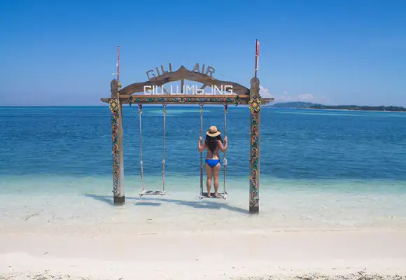 A woman in a swimsuit and hat standing on a wooden ocean swing in turquoise water at Gili Air beach.