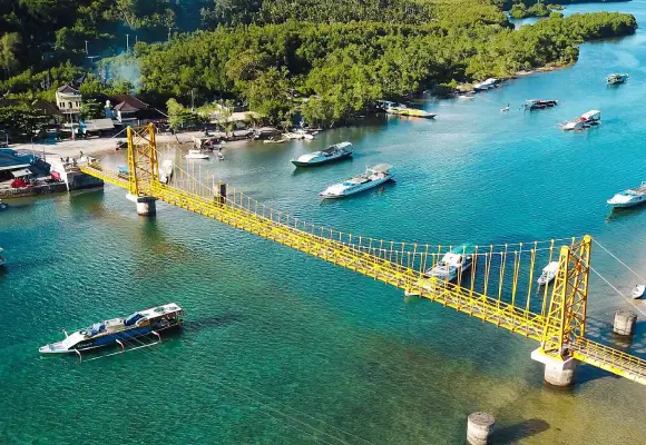Aerial view of the bright Yellow Bridge connecting Nusa Lembongan and Nusa Ceningan across clear blue water with boats anchored nearby.