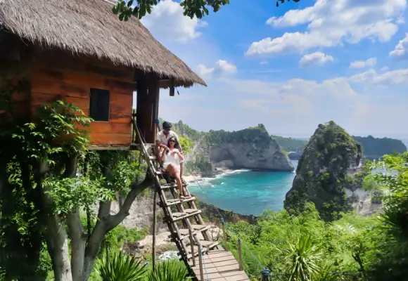 Couple sitting on the wooden stairs of the Rumah Pohon Treehouse overlooking turquoise ocean waters and limestone cliffs in Nusa Penida, Bali.